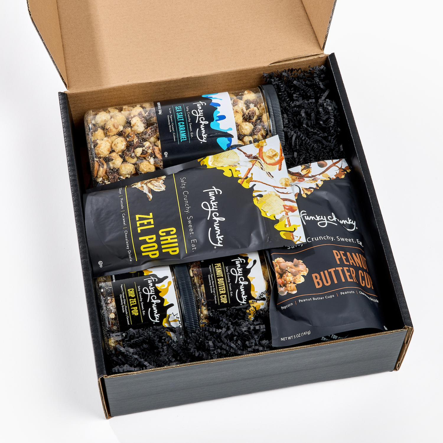 An open black gift box filled with Funky Chunky Gourmet Popcorn mix of bags and canisters - Sea Salt Caramel, Chip Zel Pop and Peanut Butter Cup arranged with black crinkle paper filler on a white background.