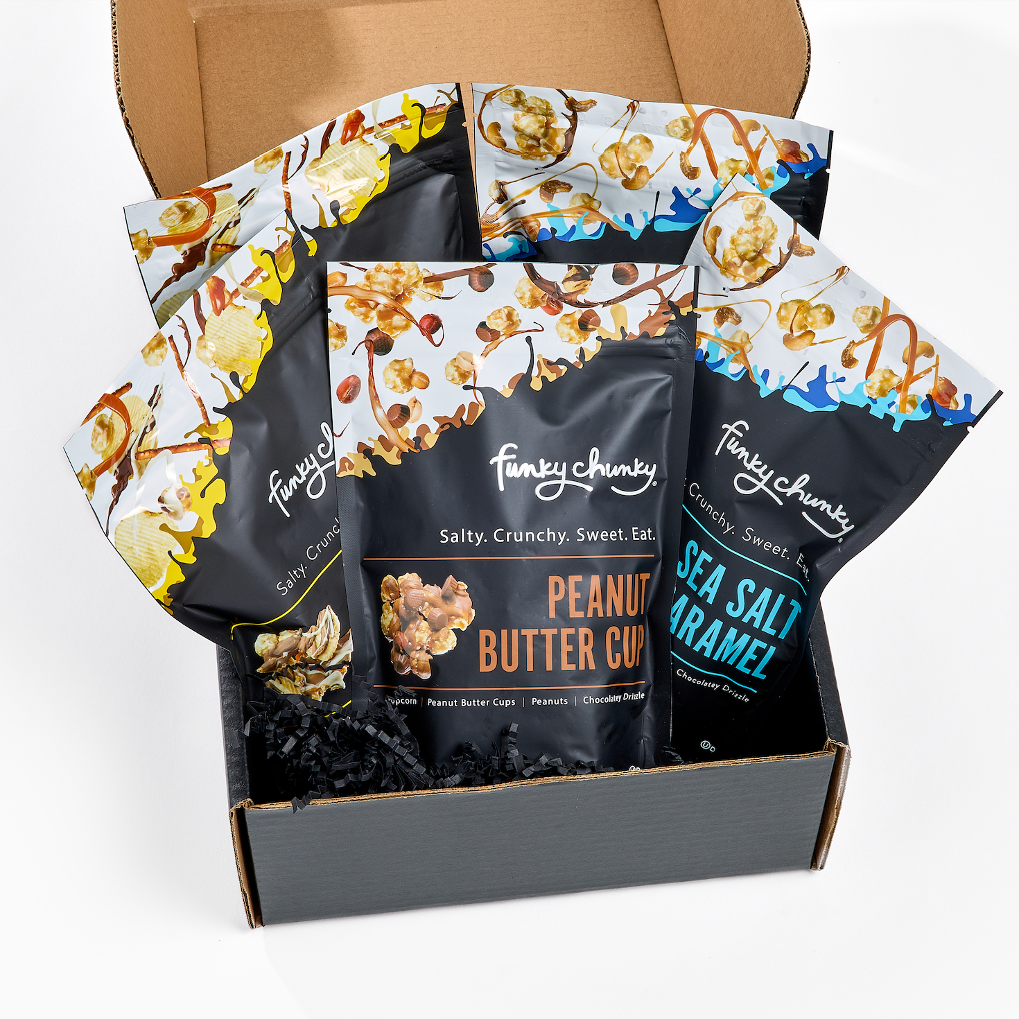 An open cardboard gift box filled with several bags of Funky Chunky gourmet popcorn mix on a clean white background. The front bag is Peanut Butter Cup, with two other bags fanned behind it - Chip Zel Pop and Sea Salt Caramel flavors and the box base is lined with black crinkle paper filler.