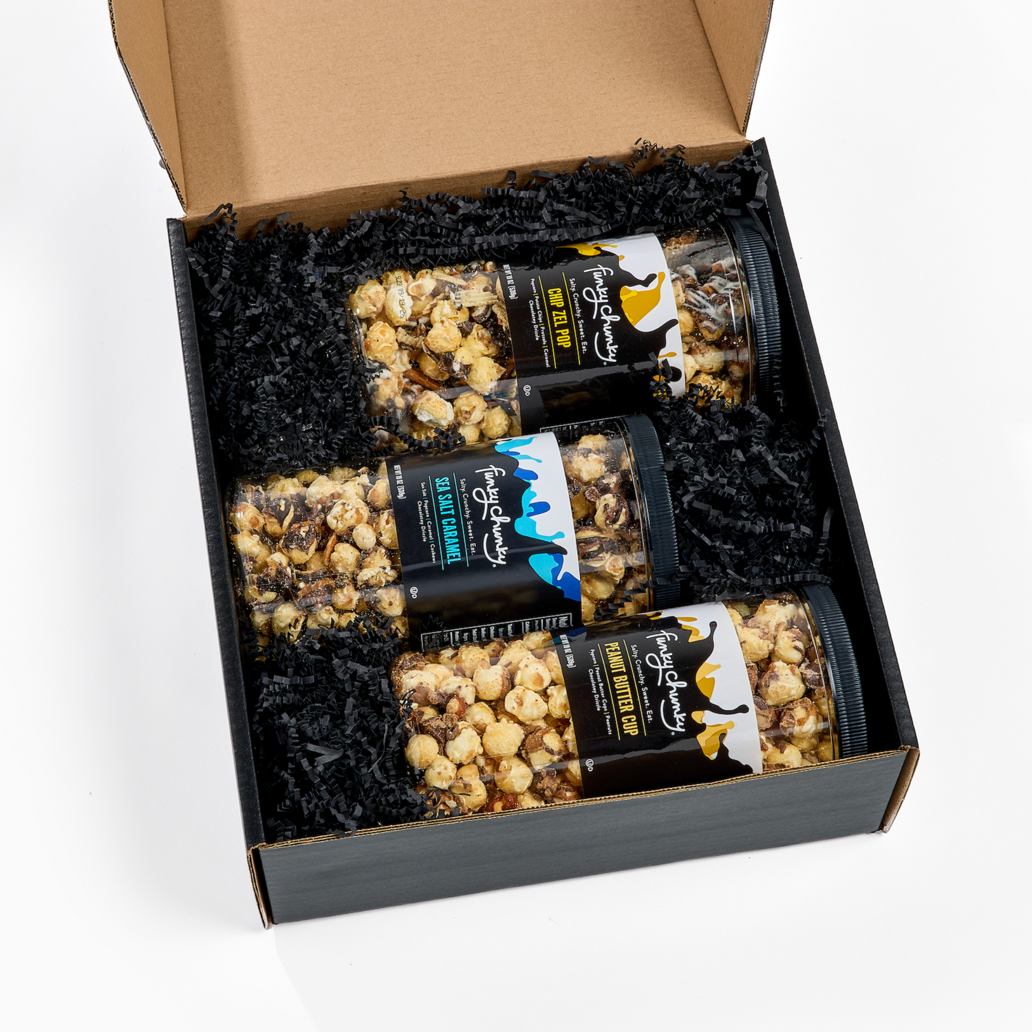 An open kraft gift box filled with black crinkle paper and four clear jars of popcorn: Funky Chunky Chip Zel Pop, Sea Salt Caramel and Peanut Butter Cup photographed on a clean white background.